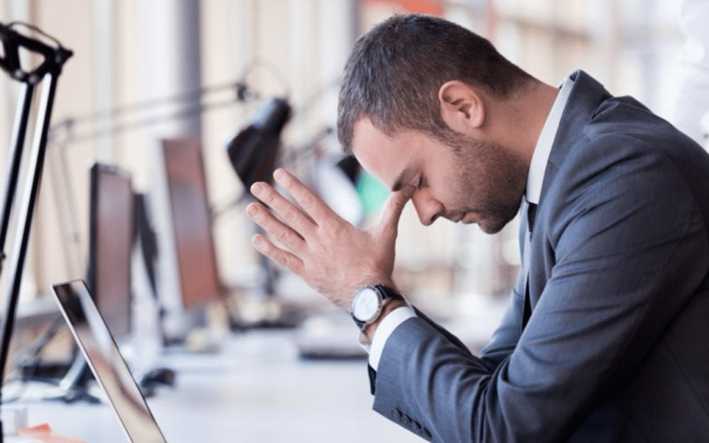 Man in a suit sits at a desk with head bowed and hands clasped, looking stressed in an office setting
